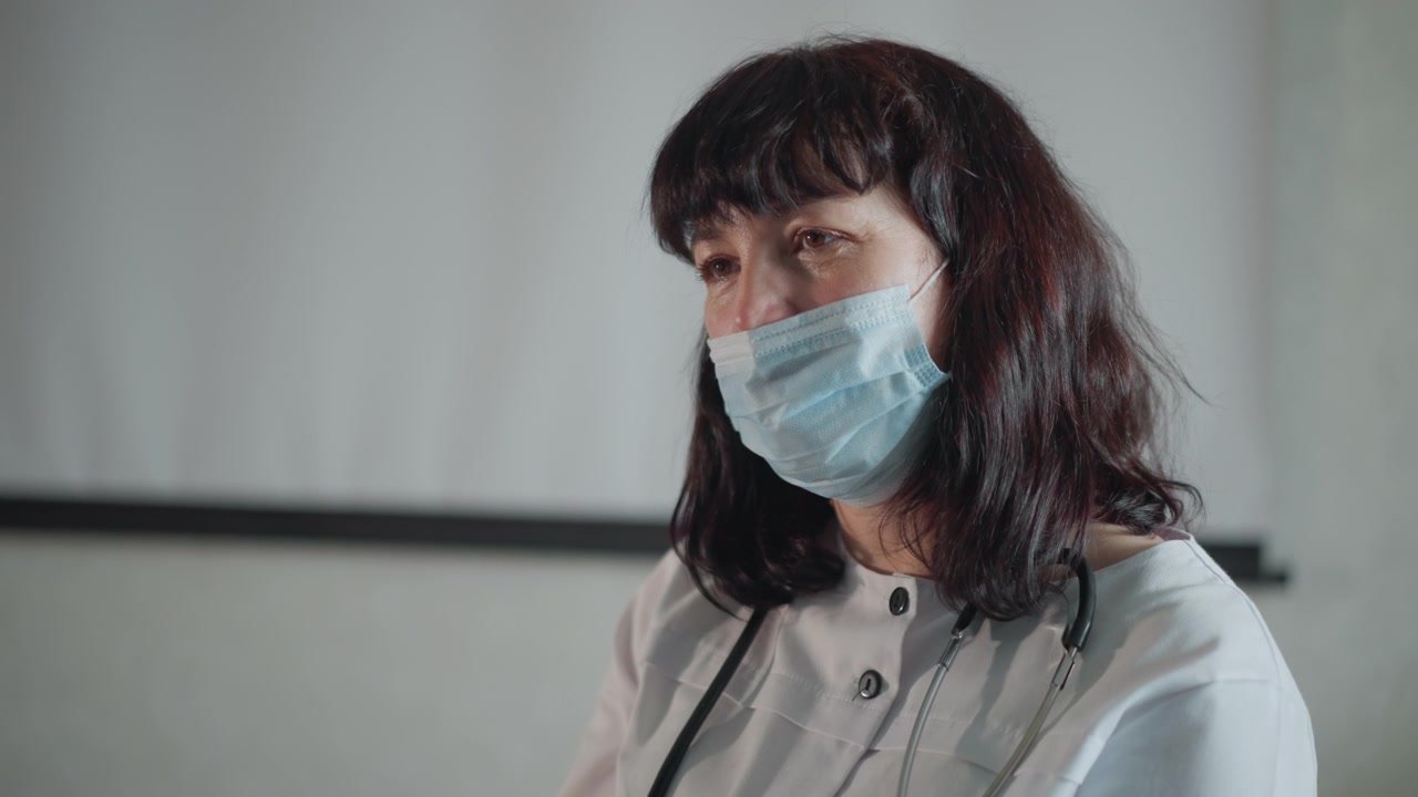 Clinica donna mascherata in sala di consultazione, ascolta attentamente con lo stetoscopio, indossa mascherina chirurgica e uniforme, espressione calma, prende appunti, si prepara per l'esame, illuminazione soffusa