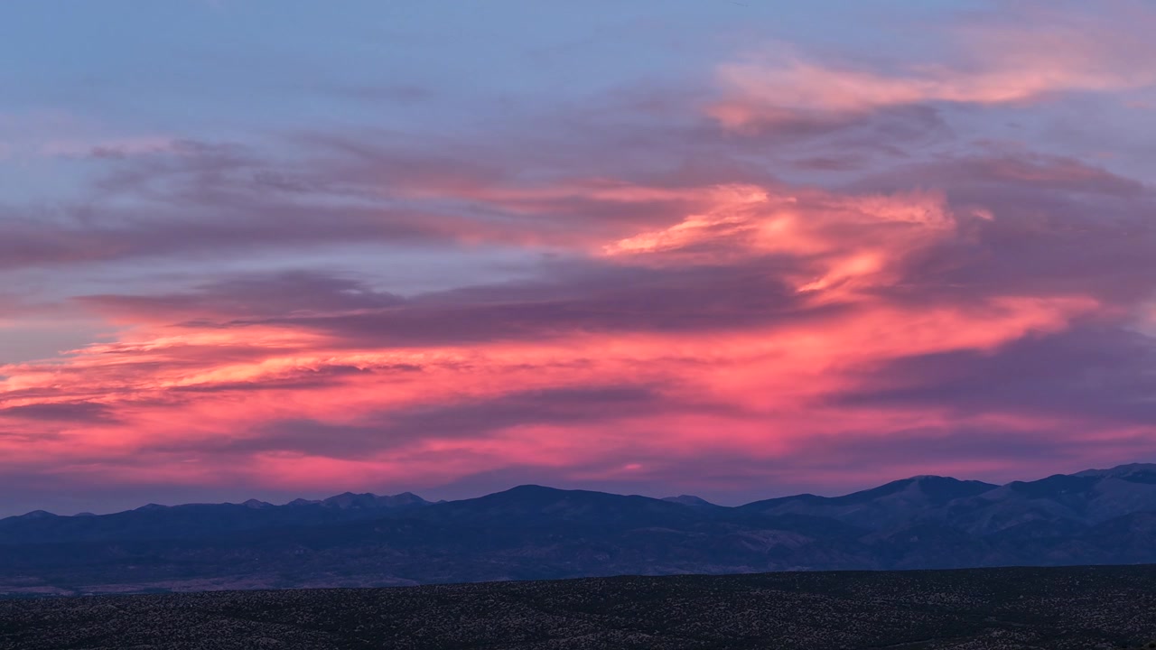 Timelapse 4K UHD che cattura un tramonto surreale e vibrante. Brillanti colori rosa neon e arancione si spostano dinamicamente attraverso uno spesso strato di nuvole che riposano sopra una scura cresta montuosa del sud-ovest.