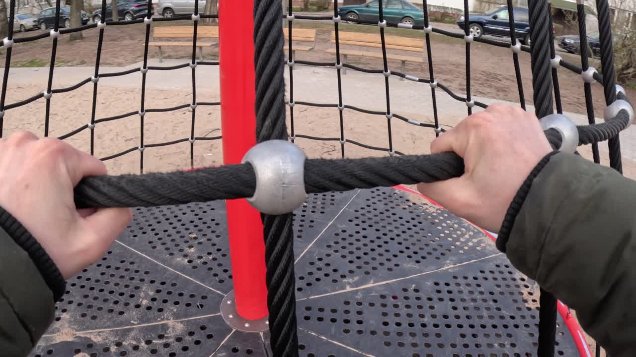 Hands Gripping Rope Net Of Revolving Climbing Tower At Playground. POV, slow motion