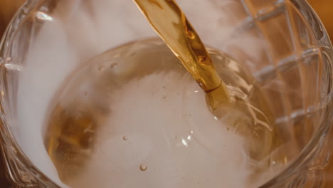 bourbon whiskey pouring over round ice cube in cocktail glass at bar in slow motion