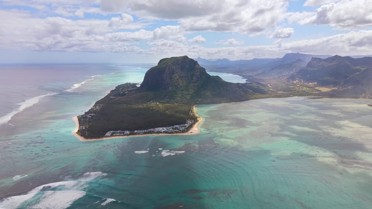 Expansive drone shot of Le Morne Brabant and the famous underwater waterfall illusion in Mauritius, surrounded by turquoise waters, coral reefs, and tropical scenery under bright clouds