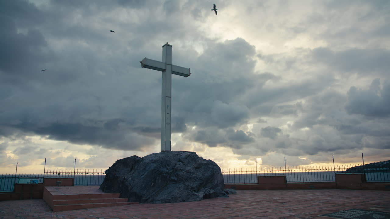 Vista grandangolare di un'alta croce bianca montata su un cumulo roccioso, stagliata contro drammatiche nuvole temporalesche e un cielo al tramonto luminoso sulla costa mediterranea