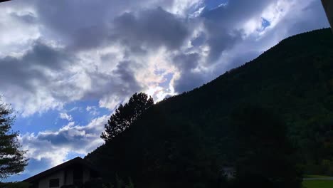 Footage-of-heavy-moving-clouds-and-mountain-with-trees,-sunlight-in-the-background-and-house-on-the-foreground