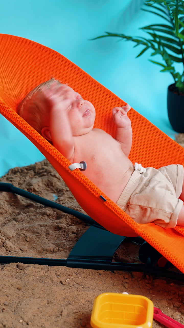 Adorable newborn in shorts yawning and stretching in the orange chair. Infant kid on the improvised beach in studio. Side view. Vertical video