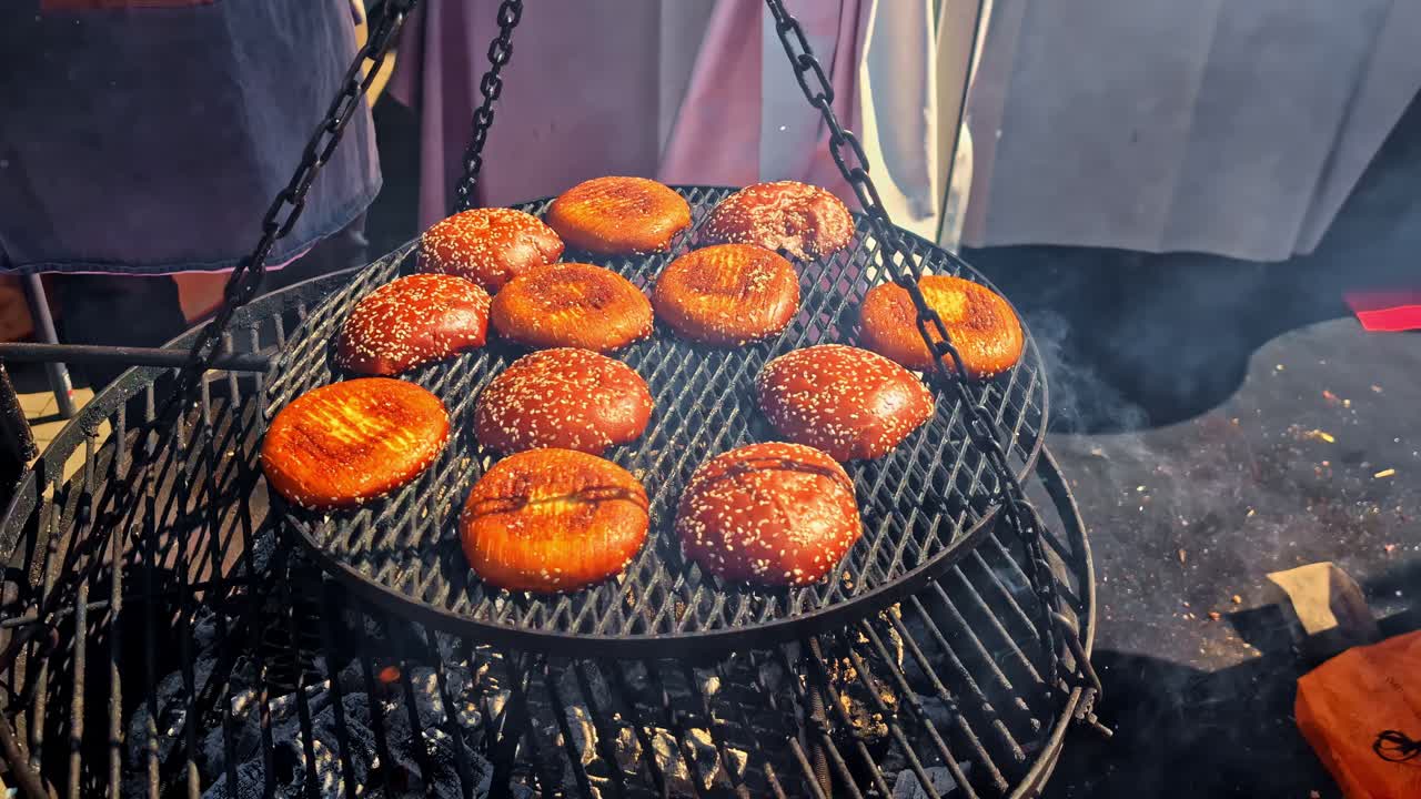 Açık ateşte ızgara yapılan, asılı bir barbekü ızgarasında yavaşça kızaran hamburger ekmeklerinin yakın çekimi. Altın sarısı ekmekler, duman ve sıcak ışık, davetkar bir açık hava pişirme sahnesi yaratıyor.