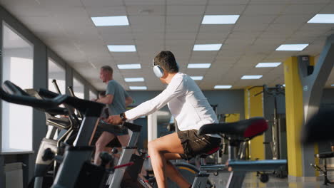 freelancer in white activewear cycles on stationary bike looking at gym activity, while runner on treadmill wipes face with towel and other equipment and people visible under bright ceiling lights