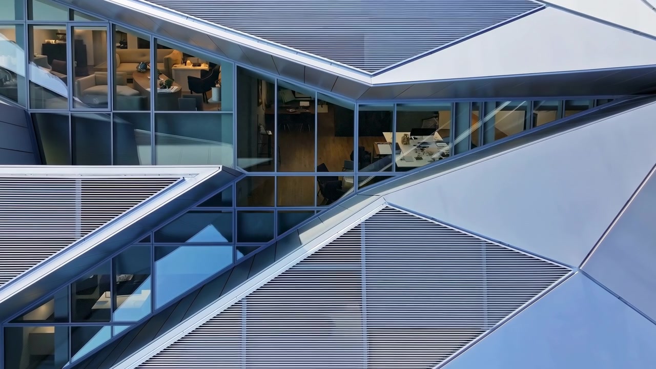 Vista aérea oblicua de la fachada de una oficina contemporánea con pasillos de cristal diagonales que se cruzan con el exterior revestido de metal. La iluminación interior cálida revela