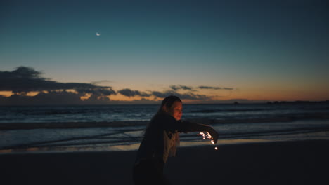 Frau-Spielt-Mit-Wunderkerze-Am-Strand-Bei-Sonnenuntergang-Und-Feiert-Silvester.-Mädchen-Hat-Spaß-Beim-Tanzen,-Schwenkt-Wunderkerzen-Feuerwerk-Und-Genießt-Die-Feier-Am-Meer