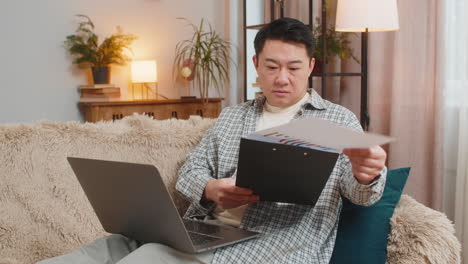 chinese male accountant using laptop working with financial graph documents reports of home income
