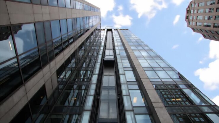 Explorando la arquitectura moderna: Una vista impresionante de un elegante edificio de cristal que se extiende hacia el cielo con reflejos y un vibrante telón de fondo urbano.