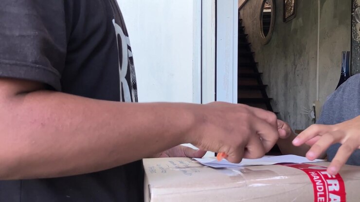 Slow motion close-up of a customer's hand signing a delivery receipt with a pen on top of a fragile package held by a courier at the doorstep. E-commerce shipping and logistics concept.