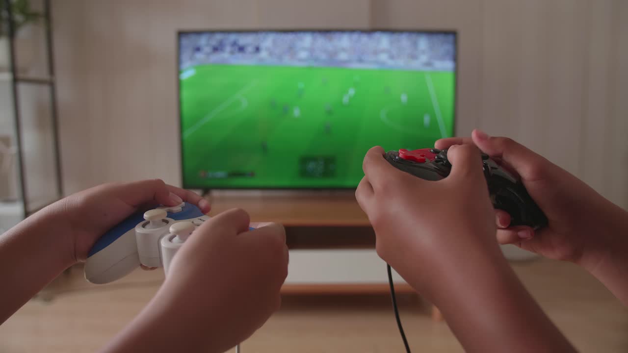 Primo piano delle mani di bambini che tengono un joystick e giocano a un videogioco di calcio in TV.