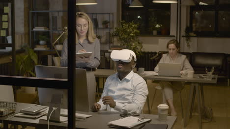 American Man Sitting At Desk In The Office Wearing Virtual Reality Glasses While A Woman Employee Is Controlling Him With Tablet 1