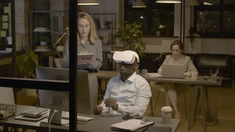 american man sitting at desk in the office wearing virtual reality glasses while a woman employee is controlling him with tablet 1