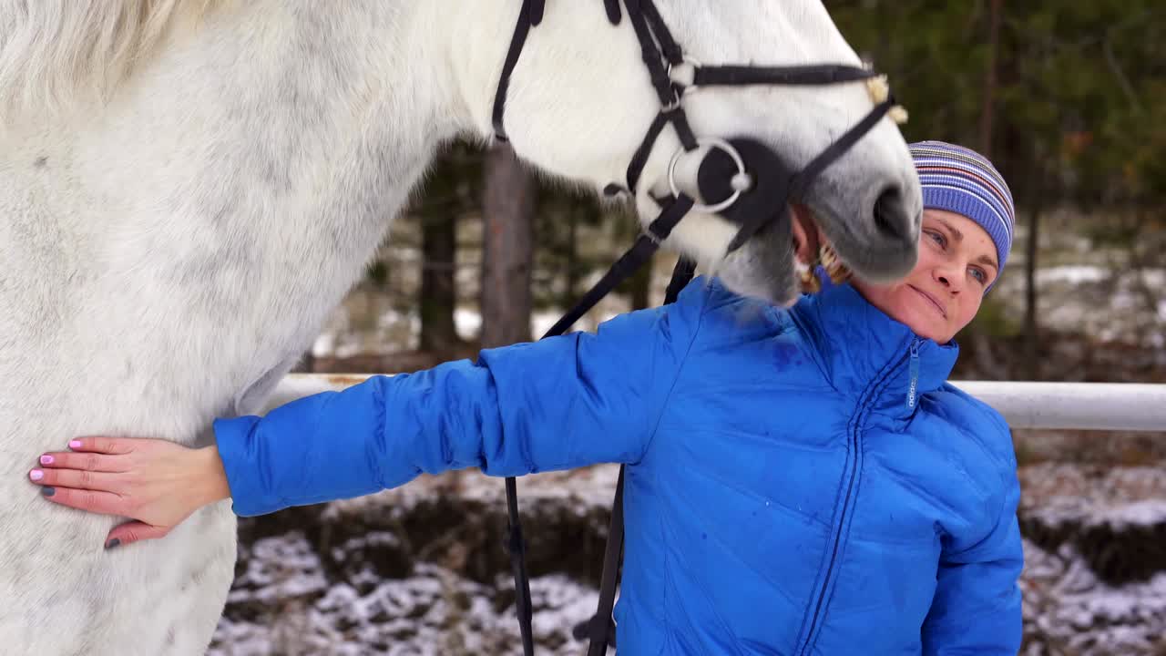 una mujer joven con una chaqueta y una gorra deportiva acaricia a un caballo blanco