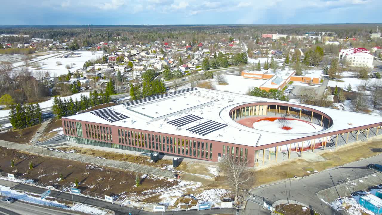 Aerial drone footage view of Saku city new school or highschool building during spring sunny day with white snow covering the modern building rooftop. Some brown grass visible and roads around.