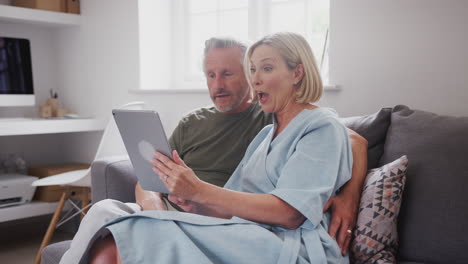 Senior-Couple-Sitting-On-Sofa-At-Home-Making-Video-Call-Using-Digital-Tablet-Together