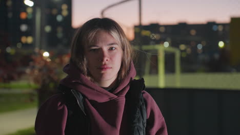 cheerful female athlete in hoodie and jumper stands outdoors under night lights gazing confidently into distance against urban fence and court at dusk with glowing city bokeh illuminating mood