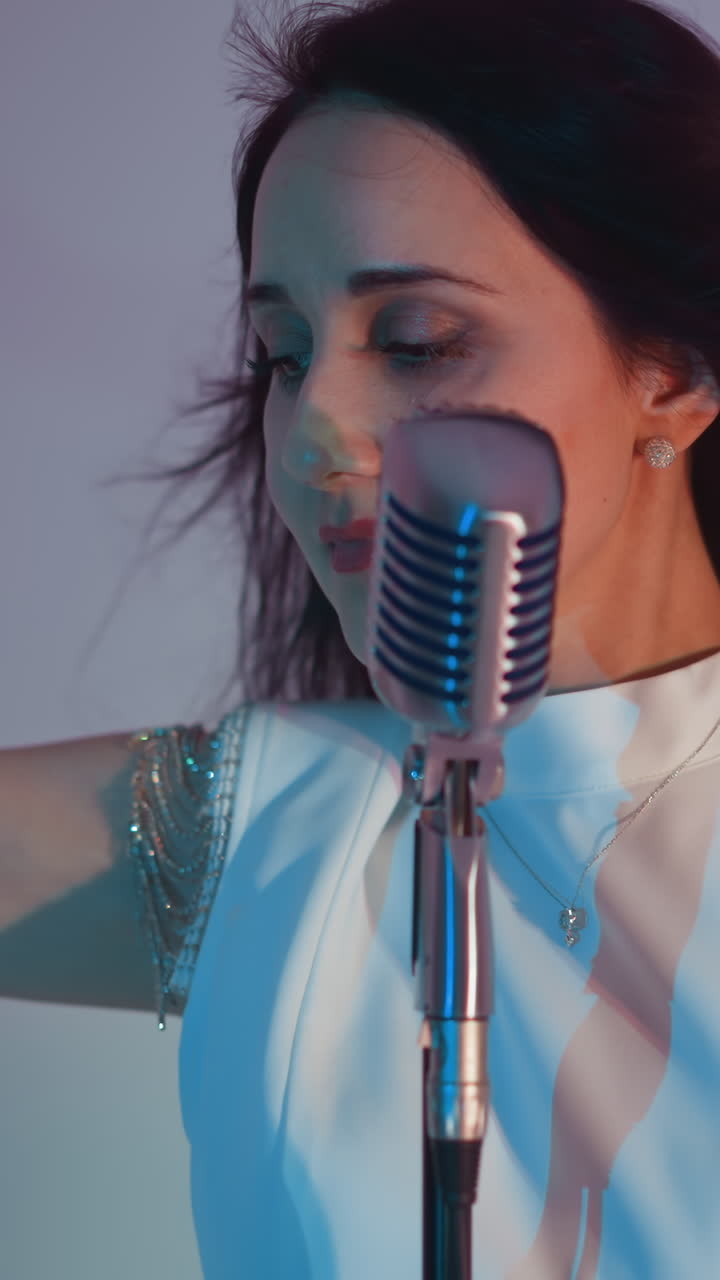 색 드레스를 입은 가수가 은 분홍색과 파란색 조명이 한 색 배경에 머리카락을 고 빈티지 마이크에 연주하는 동안 손을 공중에 우아하게 움직입니다.