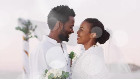 animation of light spots over happy african american bride and groom embracing on beach at wedding