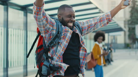 Young-African-American-cheerful-man-traveller-with-backpack-tapping-and-scrolling-on-smartphone-at-bus-stop