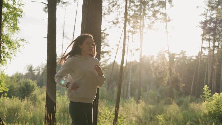 Corredora se prepara para maratona na floresta. Jovem pratica corrida para melhorar a resistência antes da competição ao pôr do sol. Rotina esportiva ao ar livre.