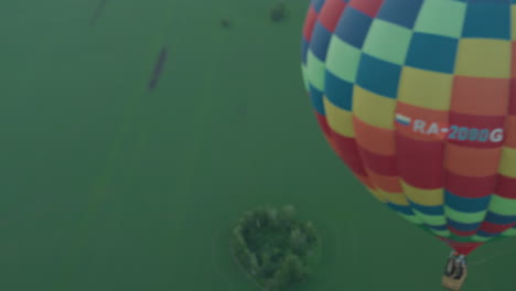 vibrant checkered hot air balloon viewed from above drift over vast green farmland at dusk, burner flame visible inside gondola, serene sky backdrop