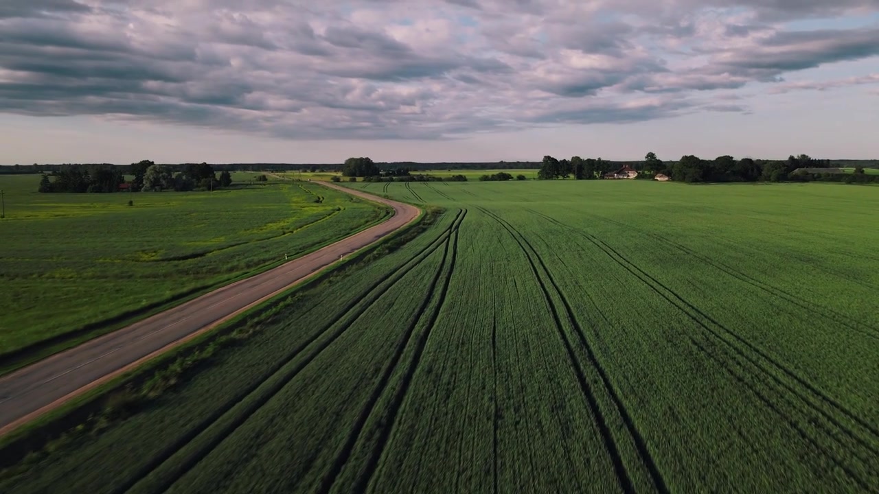 Drohnenaufnahme, die vorwärts fliegt und dabei sinkt, während Autos auf die Drohne zufahren und darunter hindurchfahren. Die Aufnahme enthüllt weiterhin die weiten Felder und die offene ländliche Landschaft unter einem bewölkten Himmel