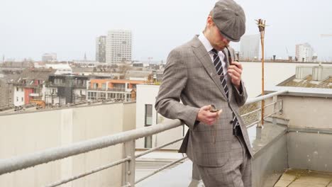 a well dressed man with a beret looking on his pocket watch while smoking an oldschool pipe on the rooftop during a foggy wintersday