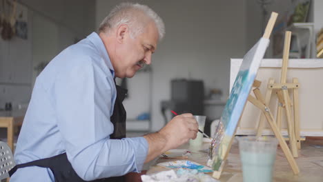 an elderly man in an apron paints a picture with a brush while sitting at the table