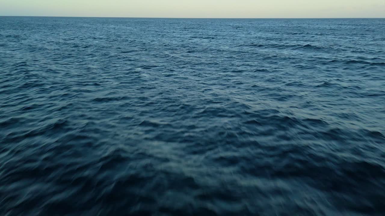 Drone tilt down to dark blue tropical ocean water with sunset glow on horizon, fade to black