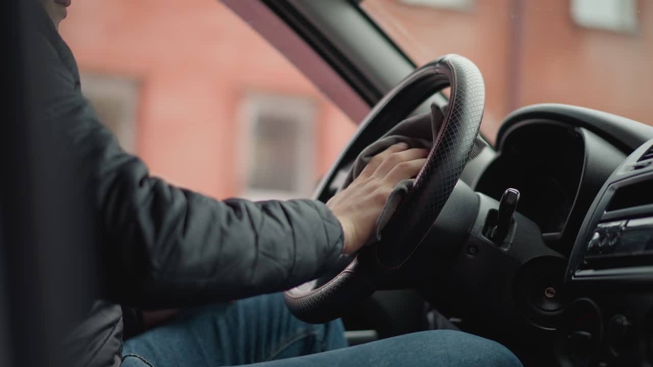 Lower view of young adult seated in car wiping leather steering wheel with handkerchief in smooth circular motion through open window highlighting careful hands focus interior details closeup vehicle