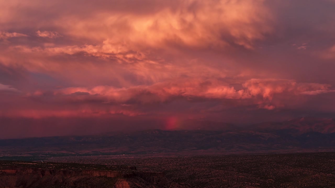 Atmosphärische 4K-Zeitrafferaufnahme einer leuchtenden Regenbogensäule, die einen majestätischen Berggipfel trifft. Dramatische Sonnenuntergangswolken und lebendige farbenfrohe Himmel entwickeln sich, während verblassende Dämmerungsschatten ein zerklüftetes Wüstental verschlingen.