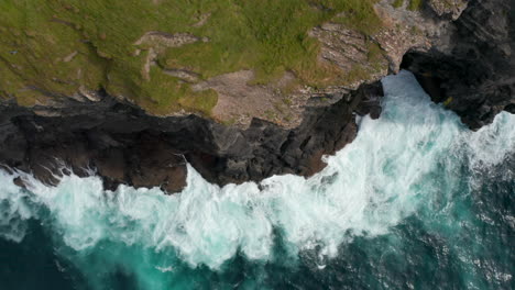 Aerial-birds-eye-overhead-top-down-ascending-view-of-rough-waves-crashing-on-rocky-coast.-Green-grass-on-top-of-cliffs-high-above-clear-water.-Kilkee-Cliff-Walk,-Ireland