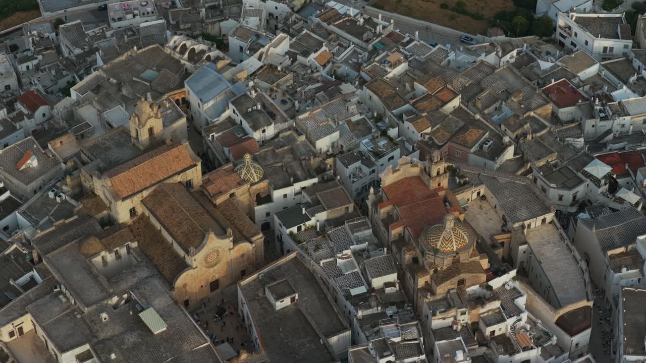 Top-down drone view of historic white city of Ostuni at dusk, showing dense buildings, rooftops, and church domes, Puglia, Italy