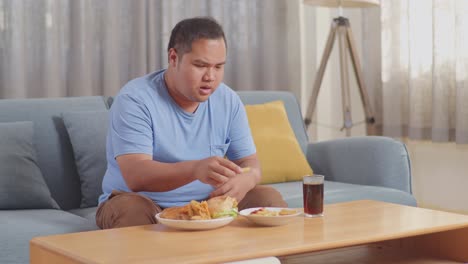 a fat asian man having a headache while dipping french fries in ketchup eating fast food on a sofa in the living room at home