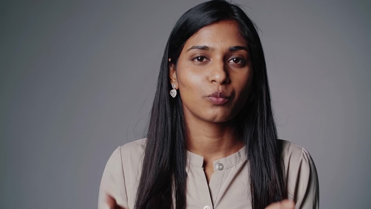 A woman speaks directly to the camera in a video, shot from a medium close-up angle, with a neutral