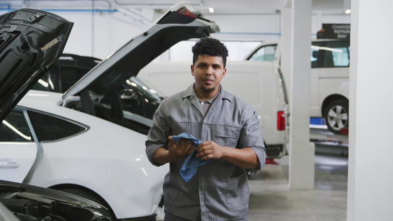 mecánico de automóviles afroamericano limpiándose las manos con un trapo y mirando a la cámara