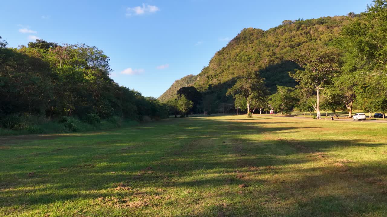 Campo tropicale illuminato dal sole con vista statica e ampia, circondato da una fitta foresta e montagne
