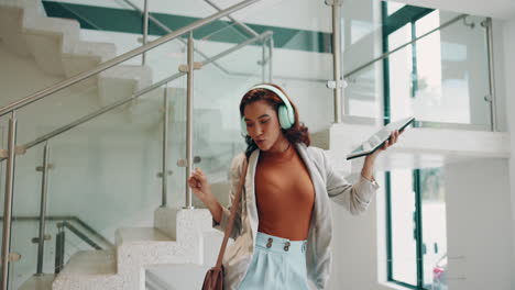 woman walking down the stairs in an office building, listening to music on her headphones and holding a tablet