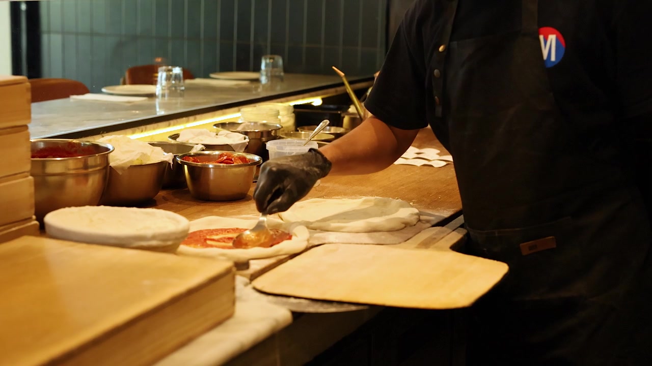 Chef con guanti neri spalma salsa di pomodoro sull'impasto della pizza in una cucina professionale.