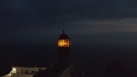 Aerial-drone-flying-forward-towards-lighthouse-shining-head-fresnel-lens-in-Lagos-Algarve,-Portugal,-dusk