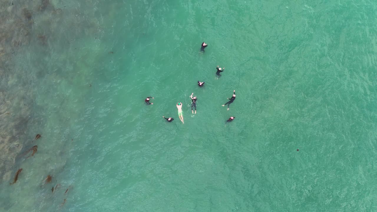Aerial footage captures swimmers practicing in turquoise waters near Port Campbell, Australia. The scene conveys teamwork and dynamic motion