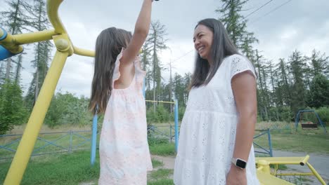 a pregnant mother and her young daughter enjoy playful time together at a playground in the park, surrounded by trees and greenery