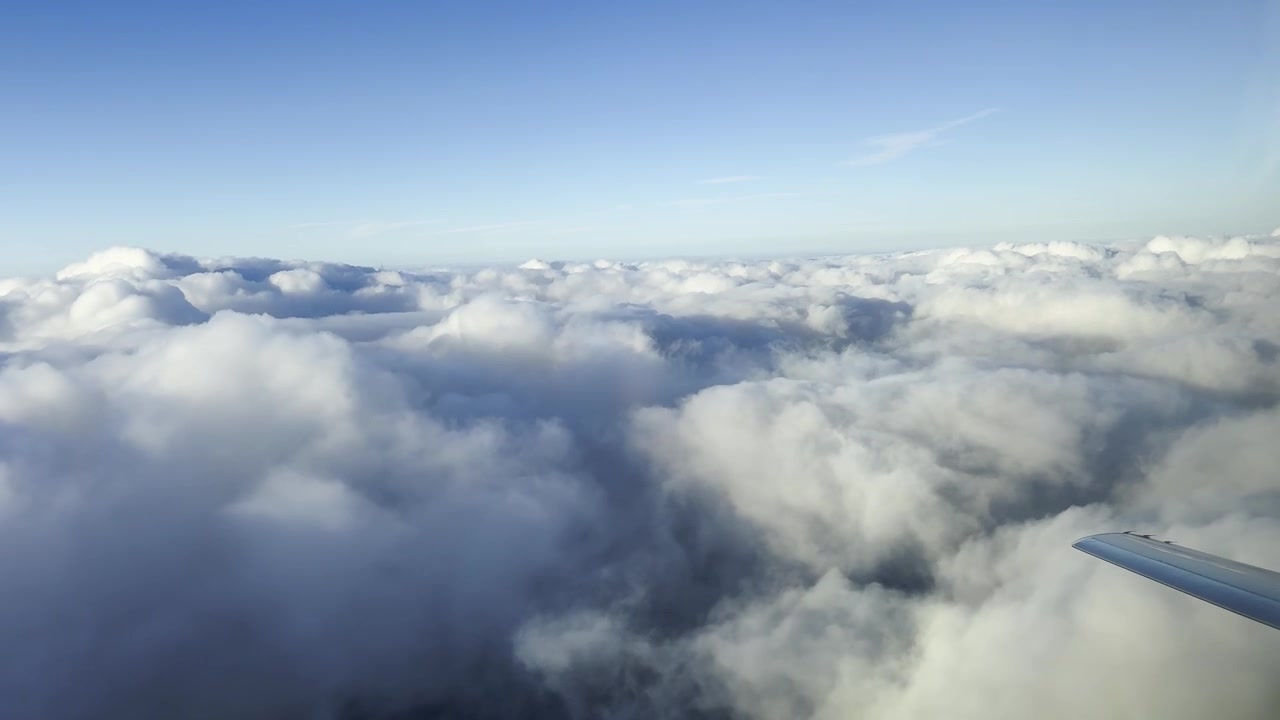 Luftfoto fra et flyvindue, der flyver over et tæt tæppe af bløde hvide cumulusskyer, med flyets vinge synlig i billedet under solnedgang.