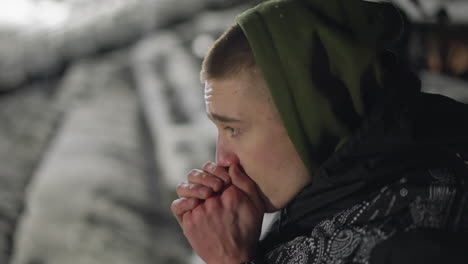 young man in winter hoodie blows air into hands to stay warm during cold night, breath visible in frosty air, soft background lights and snow covered surroundings emphasize chill environment