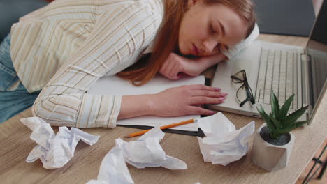 Tired-woman-at-home-office-falling-asleep-on-table-with-laptop-computer,-crumpled-sheets-of-paper