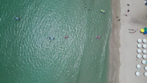 Vogelperspektive-Von-Oben-Auf-Den-Strand-Mit-Weißen-Sonnenschirmen,-Die-In-Einer-Reihe-Am-Ufer-Aufgestellt-Sind,-Andere-Bunte-Sonnenschirme-Rund-Um-Das-Meer,-Ozeanwasserwellen,-Die-Kommen-Und-Gehen,-Kleine,-Lebhafte-Boote,-Die-In-Der-Nähe-Des-Urlaubs-Oben-Vor-Anker-Liegen