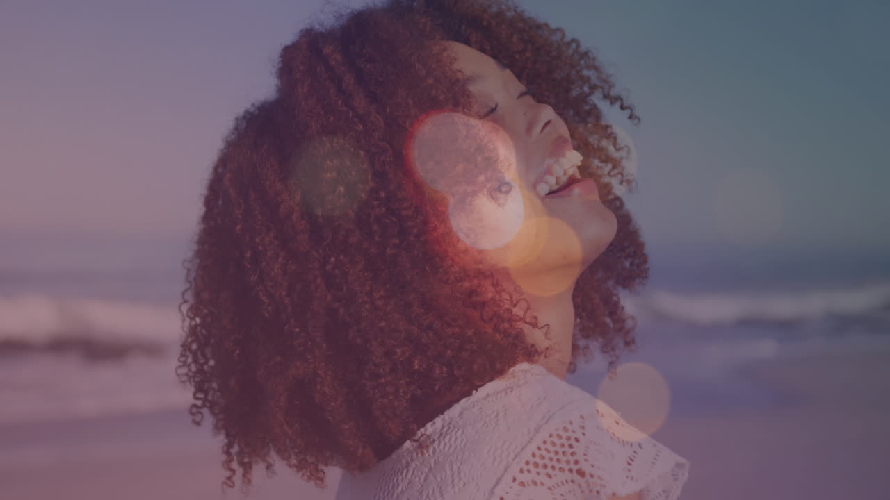 Animation of lens flares over portrait of biracial young woman with afro hair laughing at beach