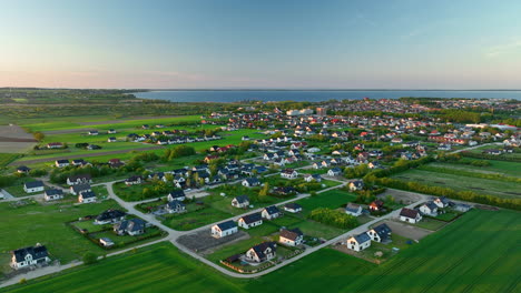 Vista-Aérea-De-Un-Pueblo-En-Puck,-Połczyno,-Con-Numerosas-Casas-Rodeadas-De-Campos-Verdes-Y-Un-Cuerpo-De-Agua-Distante-Al-Atardecer